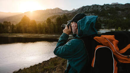 Älterer Mann auf Wandertour, der die Aussicht mit einer Digitalkamera fotografiert. Ein Wanderer steht am Fluss im Wald und macht Fotos. - JLPSF17355