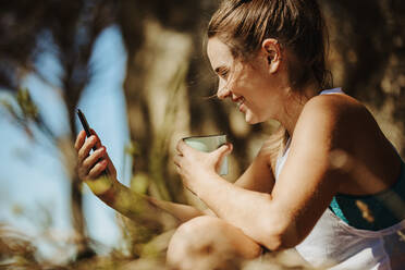 Lächelnde junge Frau schaut auf ihr Handy und trinkt einen Kaffee im Freien während einer Wanderung. Sportliche Frau macht eine Pause während des Wandertrainings. - JLPSF17360