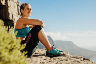 Eine fitte Frau, die auf einer Klippe sitzt, die Aussicht genießt und lächelt. Eine Wanderin, die sich auf einem Berggipfel ausruht. - JLPSF17369