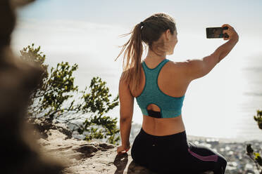 Rückansicht einer fitten jungen Frau, die auf einer Klippe sitzt und ein Selfie macht. Frau in Sportkleidung macht ein Selbstporträt mit Handy auf einem Berggipfel. - JLPSF17372