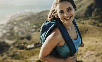 Frau mit Rucksack, die einen Berg hinaufwandert. Bergsteigerin schaut in die Kamera und lächelt, während sie einen Felsen besteigt. - JLPSF17383
