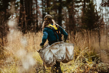 Mann arbeitet im Wald, um neue Bäume zu pflanzen, er geht durch trockenes Gras und trägt eine Tasche mit neuen Setzlingen. Mann arbeitet in der Forstwirtschaft und pflanzt Bäume in abgeholztem Land. - JLPSF17431