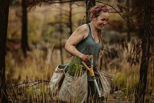 Female forest ranger planting new trees. Forester walking through dry grass carrying bag full of new seedlings and shovel. - JLPSF17455