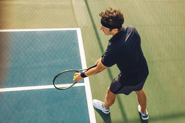 Top view of a young tennis player with racket ready to serve a tennis ball. Professional player standing at the baseline holding the tennis racket and the ball before starting the match on a hard court. - JLPSF17473