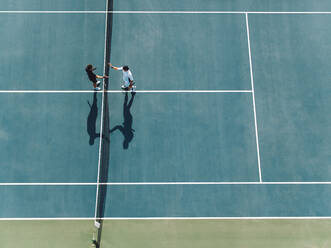 Luftaufnahme von zwei jungen Männern, die sich auf einem Hartplatz die Hände schütteln. Tennisspieler schütteln sich nach dem Spiel über dem Netz die Hände. - JLPSF17515