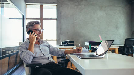 Businessman on the phone sitting at the laptop in his office. Male business professional in office talking on cell phone. - JLPSF17520