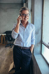 Happy entrepreneur standing by a window and talking on his cellphone. Mature businessman using phone at his office. - JLPSF17523