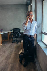 Full length shot of mature businessman talking on phone. Man standing by office window making a phone call. - JLPSF17524