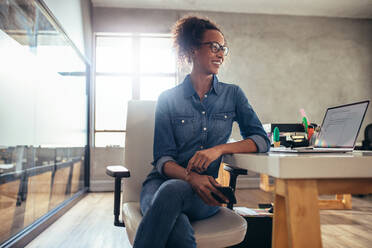 Junge Unternehmerin, die an ihrem Schreibtisch sitzt und wegschaut. Geschäftsfrau bei der Arbeit in einem kleinen Büro. - JLPSF17534