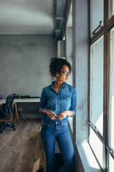 Young woman standing in office and looking outside the window. Small business owner in casuals standing by window in office. - JLPSF17539