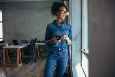 Lächelnde Frau in Freizeitkleidung, die im Büro steht und aus dem Fenster schaut. Geschäftsfrau mit Handy in der Hand, die aus dem Fenster schaut. - JLPSF17542