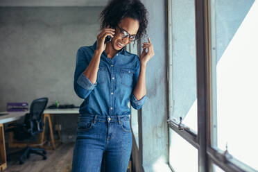 Businesswoman talking on mobile phone and smiling. Female entrepreneur standing by a window and talking over cell phone in office - JLPSF17544