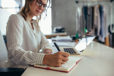 Unternehmerin schreibt in ihr Tagebuch mit Laptop auf dem Schreibtisch. Online-Unternehmerin arbeitet an ihrem Schreibtisch. - JLPSF17549