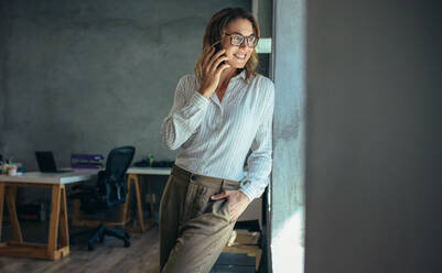 Geschäftsfrau, die mit einem Handy telefoniert und lächelt. Unternehmerin, die an einem Fenster steht und über ein Mobiltelefon im Büro spricht - JLPSF17557