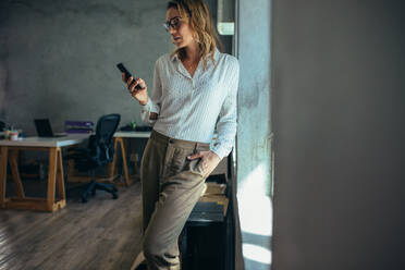 Businesswoman standing in office and using her smart phone. Female entrepreneur looking at her mobile phone. - JLPSF17558