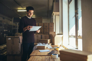 Junger Mann mit einem Papier in der Hand, der am Verpackungsbereich im Büro steht. Online Shop Besitzer arbeitet im Büro. - JLPSF17563
