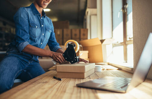 Woman packing cardboard box using tape dispenser. Female entrepreneur preparing the shipment for delivery. - JLPSF17582