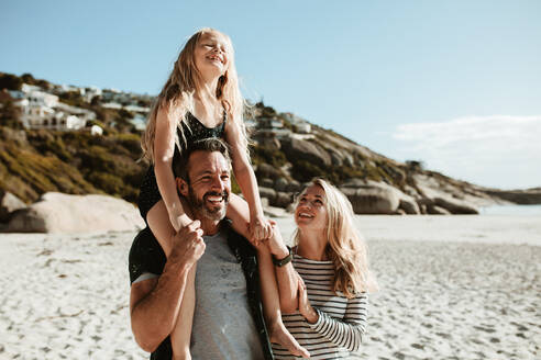 Dreiköpfige Familie im Strandurlaub: Der Vater lässt seine Tochter auf den Schultern reiten, während sie mit der Mutter am Meer spazieren gehen. - JLPSF17605