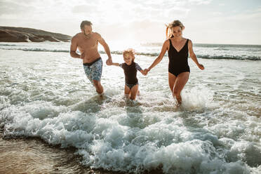 Eine dreiköpfige Familie, die sich an den Händen hält und aus dem Meerwasser läuft. Vater, Mutter und Tochter genießen ein Sommerwochenende am Strand. - JLPSF17615