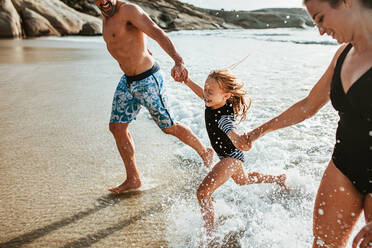 Eltern, die ihre Tochter an den Händen halten und aus dem Meerwasser am Strand laufen. Eine Familie, die ein Sommerwochenende am Strand genießt. - JLPSF17616