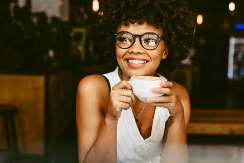 Beautiful young woman sitting at cafe with a cup of coffee and looking away smiling. African female having coffee at coffeeshop. - JLPSF17625