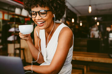 Junge afrikanische Frau, die in einem Café Kaffee trinkt und einen Laptop benutzt. Lächelnde Frau, die im Café sitzt, Kaffee trinkt und auf den Laptop auf dem Tisch schaut. - JLPSF17628