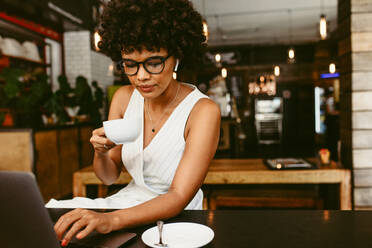 Junge Frau sitzt in einem Café, arbeitet an einem Laptop und trinkt Kaffee. Afrikanische Frau trinkt Kaffee und arbeitet an einem Laptop in einem Café. - JLPSF17630