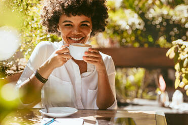 Fröhliche junge afrikanische Frau mit einer Tasse Kaffee im Café sitzend. Frau trinkt Kaffee im Coffeeshop. - JLPSF17635
