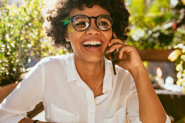 Fröhliche Frau, die in einem Café im Freien sitzt und über ein Mobiltelefon spricht. Afrikanische Frau, die in einem Café ein Gespräch am Telefon führt. - JLPSF17638