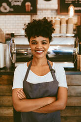 Confident female barista standing in front of a cafe counter. Woman cafe owner in apron looking at camera and smiling with her arms crossed. - JLPSF17644