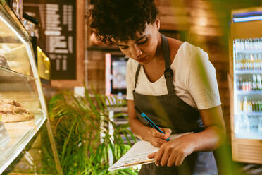 Afrikanische Frau, die in ihrem Café arbeitet, den Vorrat überprüft und sich Notizen macht, während sie die Vitrine mit köstlichen Desserts betrachtet. - JLPSF17645