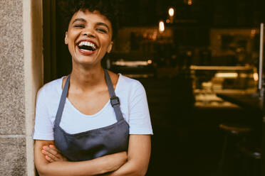 Cheerful female cafe owner standing at the door with her arms crossed. African woman in apron standing with her arms crossed and laughing. - JLPSF17648