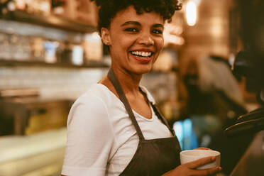 Weibliche Barista hält eine Tasse neben einer Kaffeemaschine. Weibliche Caféangestellte bereitet Kaffee in der Maschine zu. - JLPSF17651