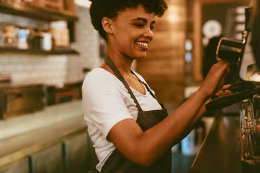 Afrikanische Barista-Frau, die eine Kaffeemaschine benutzt, um eine schöne Tasse Kaffee zuzubereiten. Café-Mitarbeiter, der einen Kaffee zubereitet. - JLPSF17652