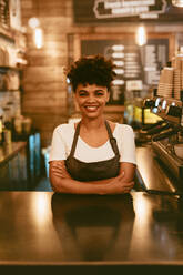 Lächelnde afrikanische Frau in Schürze, die an der Theke eines Cafés steht. Selbstbewusste Barista-Frau, die hinter der Theke steht. - JLPSF17653