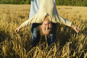 Playful girl with arms outstretched hanging upside down in front of father at field - SEAF01441