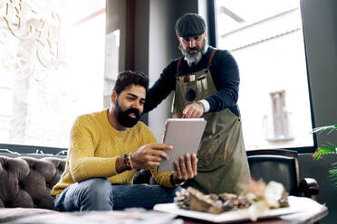 Niedriger Winkel des ethnischen männlichen Kunden, der ein Tablet benutzt und eine Frisur auswählt, während der Friseur in der Nähe des Sofas im Friseursalon steht - ADSF39925