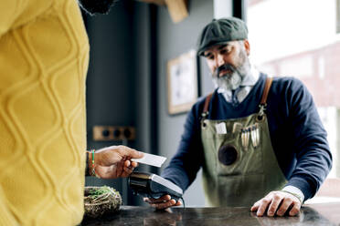 Ein männlicher Kunde bezahlt eine Frisur mit einer Plastikkarte auf einem NFC-Terminal in den Händen eines Friseurs in einem Friseursalon - ADSF39927