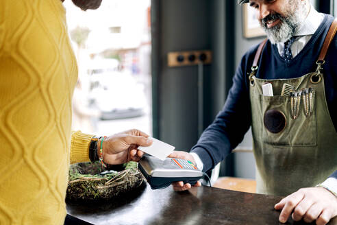 Ein männlicher Kunde bezahlt eine Frisur mit einer Plastikkarte auf einem NFC-Terminal in den Händen eines Friseurs in einem Friseursalon - ADSF39928