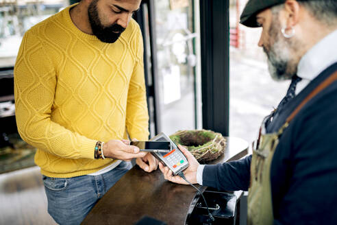 Ethnischer männlicher Kunde beim kontaktlosen Bezahlen mit dem Smartphone am POS-Terminal in einem modernen Friseursalon - ADSF39929