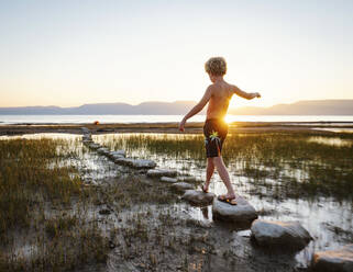 Rear view of boy (10-11) walking on stepping stones in lake at sunrise - TETF01823