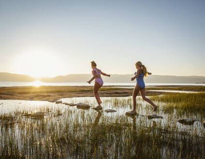 Schwestern (12-13, 14-15), die bei Sonnenaufgang auf Trittsteinen im See laufen - TETF01825