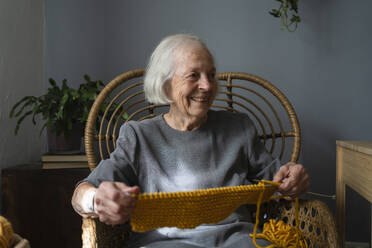 Glückliche ältere Frau mit Strickwolle und Nadel auf einem Stuhl zu Hause - SVKF00658