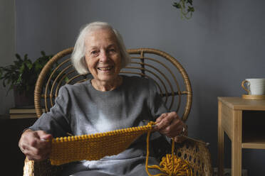 Lächelnde ältere Frau mit Strickwolle und Nadel auf einem Stuhl zu Hause - SVKF00659