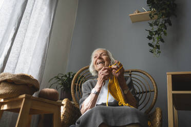 Glückliche ältere Frau strickt auf einem Stuhl im Wohnzimmer - SVKF00663