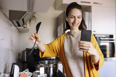 Lächelnde junge Frau, die einen Holzlöffel hält und einen Videoanruf über ein Mobiltelefon in der Küche führt - VIVF00084