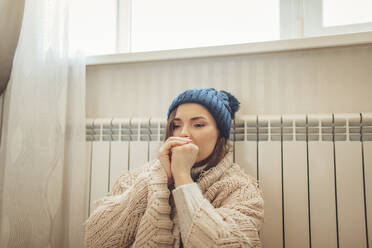 Frau mit Strickmütze, eingewickelt in eine Decke, an einen Heizkörper gelehnt - MDOF00128