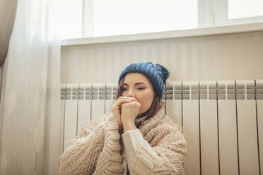 Woman wearing knit hat wrapped in blanket leaning on radiator - MDOF00128