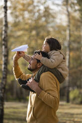 Father carrying boy holding paper airplane on shoulders in park - ANAF00255