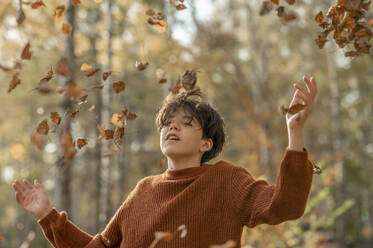 Verspielter Junge mit geschlossenen Augen wirft trockene Blätter im Park - ANAF00263